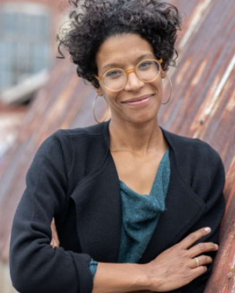 woman leaning against a wall wearing a blue top and black jacket and glasses